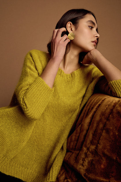 A woman in a Wide Neck Mohair Knit sweater and large round earrings sits on a brown sofa, resting her chin on her hand and gazing to the side.