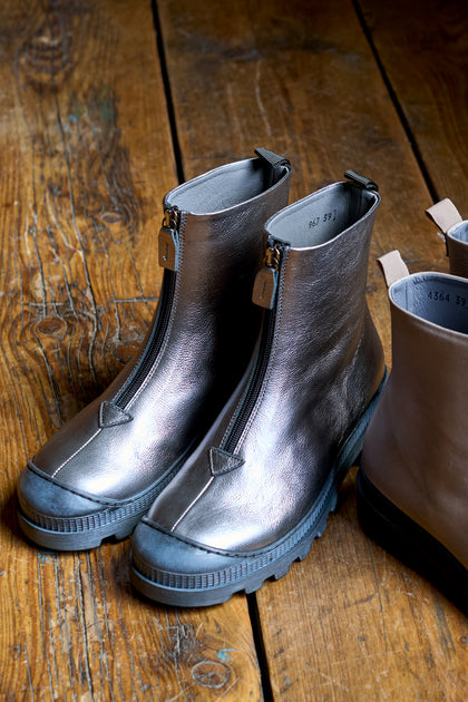 A pair of Gunmetal Zip Front Leather Boots with thick soles sits on a wooden floor, handcrafted in Italy by Lofina. A partial view of another stylish pair is also visible nearby.